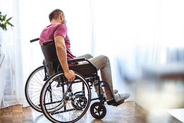 Fototapeta na wymiar Disabled Person Sits in Wheelchair Against Window. Serious Sad Caucasian Man Wearing Casual Clothes and Look at Large Panoramic View in Bright Modern Living Room or Hospital.