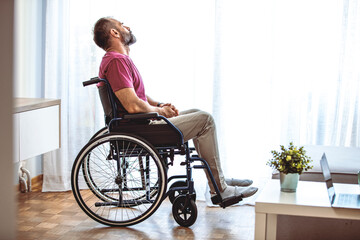 Handicapped person sitting in wheelchair and thinking about life, depression. Disabled mature man sitting on wheelchair and looking out the window in the living room at home
