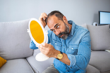 Middle-aged man concerned by hair loss. Senior man and hair loss issue. Middle aged man with alopecia looking at mirror, hair loss concept. Bearded mid adult man with alopecia looking at mirror
