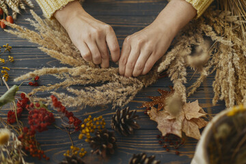 Hands holding dry grass and making stylish autumn boho wreath with wildflowers and herbs on rustic...