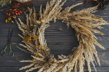 Stylish autumn rustic wreath flat lay. Making creative boho wreath with dry grass, tansy wildflowers, wheat, dog-rose berries, scissors and thread on dark wooden table. Holiday workshop