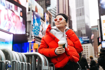 Trendy young girl over urban downtown. Stylish female hold paper coffee cup wearing fashionable eyewear and modern trend sport style clothes over New York streets background walk on Time Square in NYC