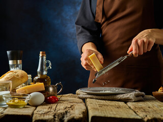 The chef grates Parmesan cheese for the classic Caesar salad. To his left are the ingredients for this delicious and healthy salad. Close-up. Careful viewing. Dark blue background.