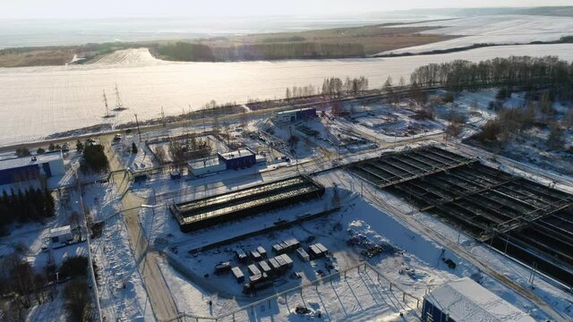 Set of basins and electricity production substation at wastewater treatment plant near frozen river at winter sunset aerial view