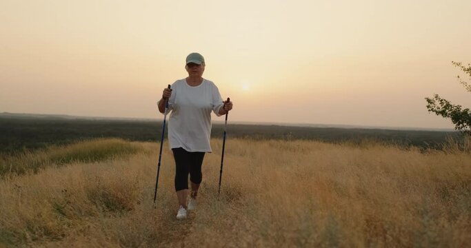 An elderly woman with nordic walking sticks walks forward through a scenic spot