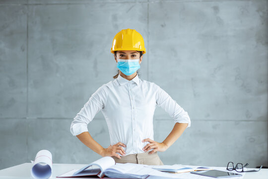 Female architect with face mask and helmet standing with hands on hips. There are blueprints and documentation on table. - Powered by Adobe