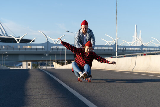 Trendy couple on longboard: hipster man teaching girl to balance on skateboard pushing her back down empty road. Happy young loving male and female spend time together with cheerful outdoor activity
