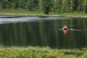 A fisherman on an inflatable rubber boat floats on a mountain lake. Life jacket orange. Beautiful landscape on a summer day.