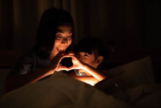 Asian Parent Bedtime With Kid, Mom Playing With Child, Reading A Book Before Sleeping In Bedroom. Mother And Daughter Enjoying On The Bed, Happy, Smiling