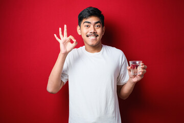 Young handsome asian man drinking glass of water pointing thumb up to the side smiling happy with open mouth