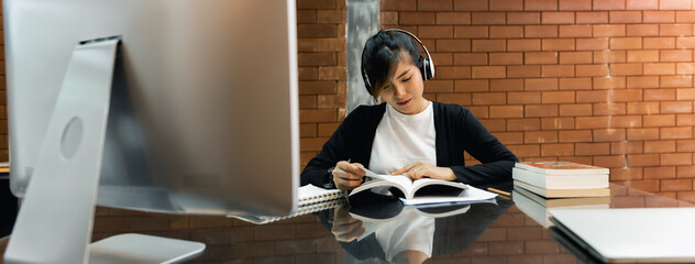 Asian scholarship student reading book on desk while wear headphone for listen teaching record and watch teacher with computer. Education research and self-improvement. Banner size with copy space.