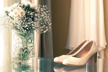 beautiful wedding slippers on a glass table and a floral arrangement with white flowers and a wedding dress in the background on the wall