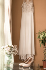 vertical shot of a beautiful wedding slippers on a glass table and a floral arrangement with white flowers and a wedding dress in the background on the wall