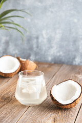 A glass of fresh, organic coconut water, milk on a wooden table under palm leaves. Chopped juicy coconuts lie nearby. Vertical photo