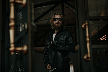 Fototapeta na wymiar Brutal caucasian man with long beard hipster in black leather jacket stay next to glass door 