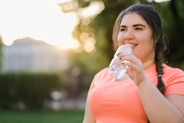 Water detox. Body refreshment. Diet nutrition. Body positive. Happy young overweight obese woman drinking from bottle in defocused copy space sunset park. - obrazy, fototapety, plakaty