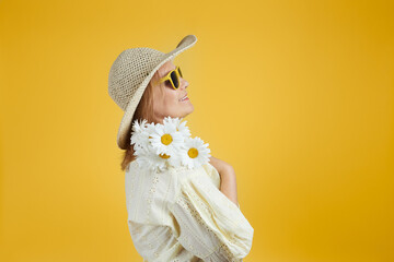 Mature woman in straw hat and sunglasses holds a bouquet of daisies. Isolate on bright yellow background