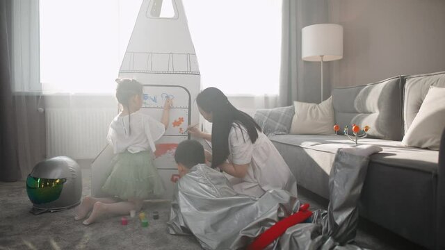 An Asian female with kids play in the living room at home, a boy in an astronaut costume lying on the floor with her mother and sister, children together with their mother paint on a cardboard model