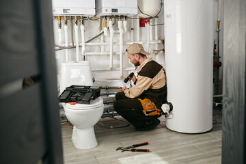 Young repairman in uniform examining part of pipe in toilet
