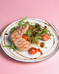 Close up still life of a tuna steak with fresh vegetables and arugula with seasoning over pastel pink background.