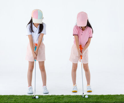 Isolated Studio Shot Of Two Pretty Cute Little Asian Golfer Sister In Sport Athlete Uniform Standing Together Holding Putter Ready To Putting Golf Ball On Green Grass In Front Of White Background