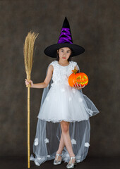 Portrait studio shot of little cute Asian kid in white witch dress costume with black purple high hat standing holding carved orange pumpkin face and magic broomstick in front of dark background