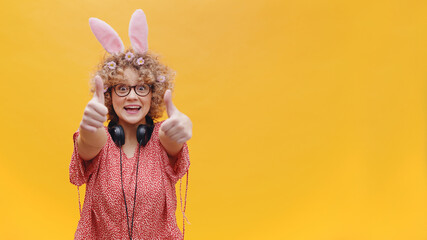 Astonished young girl wearing cute bunny ears and spectacles in a joyful mood. Headphones around her neck. Girl smiling pointing at the camera. Bright yellow background. Studio shot.
