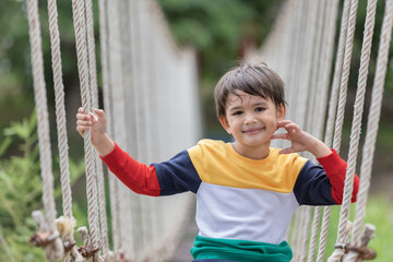 A boy wearing bright colored clothes sitting alone on rope bridge at extreme sport in adventure park.