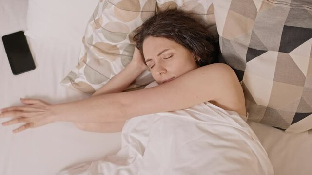 Top view of dark-haired Caucasian woman sleeping in bedroom, trying groping with hand and eyes closed her cellphone on bed, then frowning and getting back to sleep