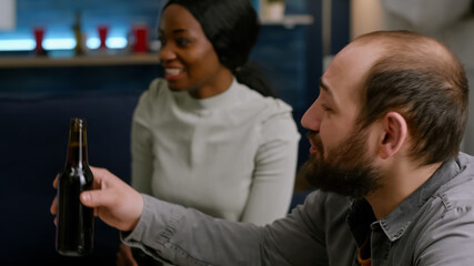 Mixed race friends cheering bottles of beer spending time together while relaxing on couch. Group of multi-ethnic people hanging out, socializing during wekeend party late at night
