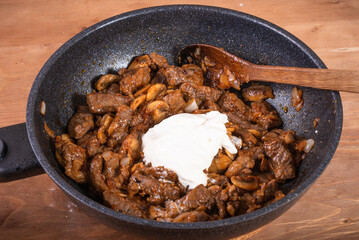 Cooking beef stroganoff in a pan - adding sour cream to a pan with fried meat and mushrooms.