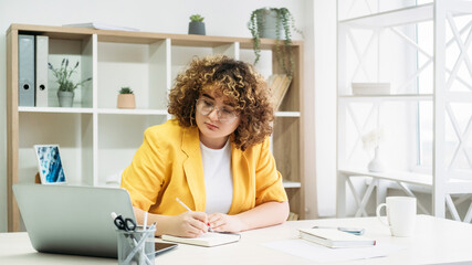Video conference. Webinar training. Remote education. Focused overweight woman in eyeglasses with curly hair studying on laptop writing notes at modern light home interior.