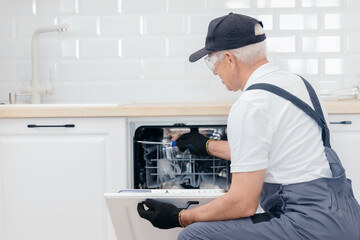 Worker man in uniform installs appliances dishwasher fittings assembling furniture in kitchen