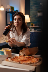 Woman holding beer bottle relaxing on couch in living room watching documentary series on television eating tasty snack. Caucasian female enjoying takeaway food home delivery at night