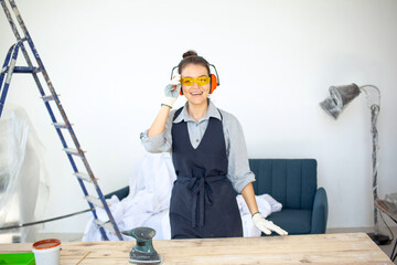 Young dark-haired woman in glasses smiling going to operate power tools in workshop preparing wooden table surface to painting