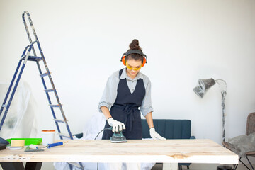 Young dark-haired woman in glasses and headphones smiling running power tools in workshop preparing wooden table surface to painting