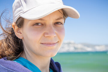 Natural face portrait of a young caucasian woman, no retouching skin, no color correction. Close-up girl outdoor portrait on a sea background, brunette in baseball cap, naturalness