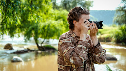 A young man with curly hair taking shots using camera in the nature, river and greenery nearby