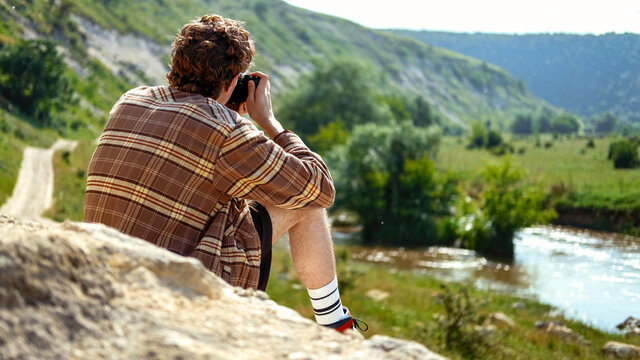 A young man with curly hair taking shots using camera in the nature, sitting on a rock, greenery and river nearby