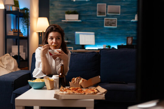 Cheerful woman eating tasty chinese food relaxing on couch during junk-food home delivered. Smiling caucasian female enjoying takeaway delivery fastfood meal in evening