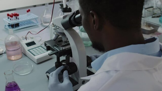 Handheld slowmo over shoulder shot of black male scientist in white coat and gloves looking into microscope while working in lab
