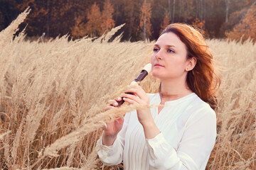 A young woman plays a flute standing in a field near forest trees