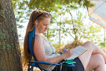 Young woman with long red hair is studying remotely online. She writes notes while watching a...