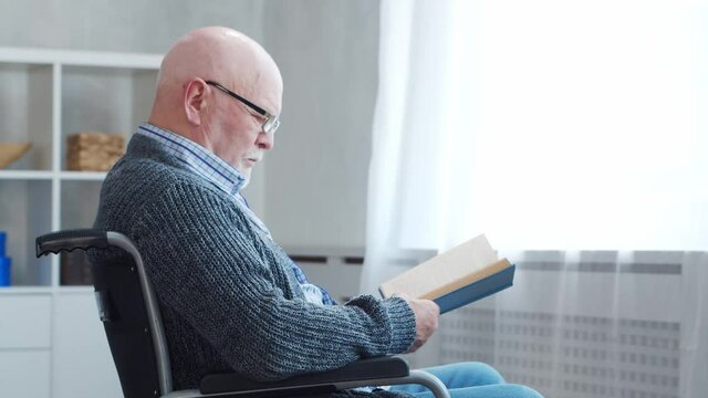 Disabled old man is sitting in a wheelchair at home alone. A handicapped person in a wheelchair is reading the book. Paralysis and disability.