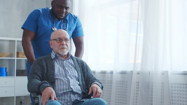African-American caregiver and old disabled man in a wheelchair. Professional nurse and handicapped patient in a nursing home. Assistance, rehabilitation and health care.