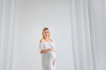 A young pregnant woman in anticipation of a baby in a white dress, hugs belly. Fashion girl in a minimalistic, isolated interior of a cut of airy fabrics. Easy, happy maternity pregnancy, surrogacy.