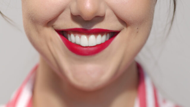 Lips with deep saturated red lipstick close-up. Caucasian brunette girl smiling wide showing crystal white teeth. Anonymous lady studio shot high quality photo image.