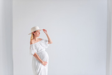 Young pregnant woman in anticipation of a baby in a white hat, dress, hugs her belly. Tender blonde girl in a minimalistic, isolated interior of airy fabrics. Happy maternity pregnancy, surrogacy