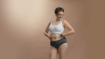 Chubby female in white bra and black briefs smiles, looks to the camera and adores her forms. Hands on the waist. Studio cowboy shot photo image.