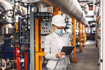 Dedicated female blond supervisor with face mask standing in heating plant next to dashboard and holding tablet to check on machinery during corona pandemic.